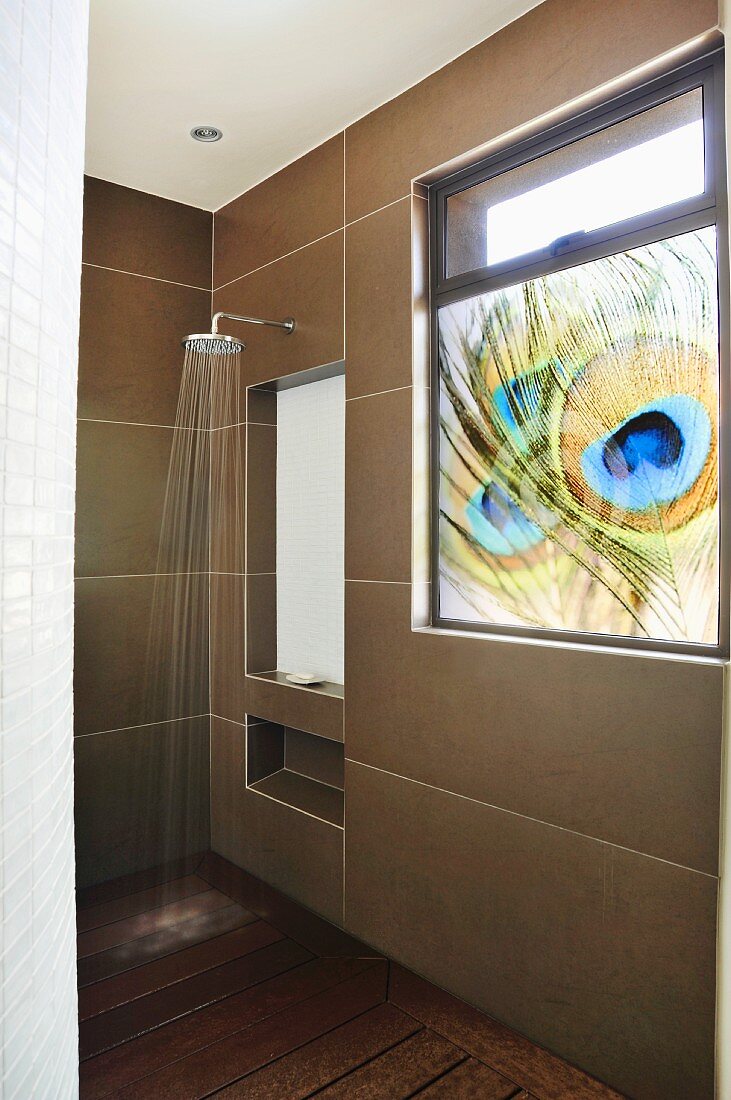 Shower head on wall with large tiles and opaque window with peacock feather motif in shower area