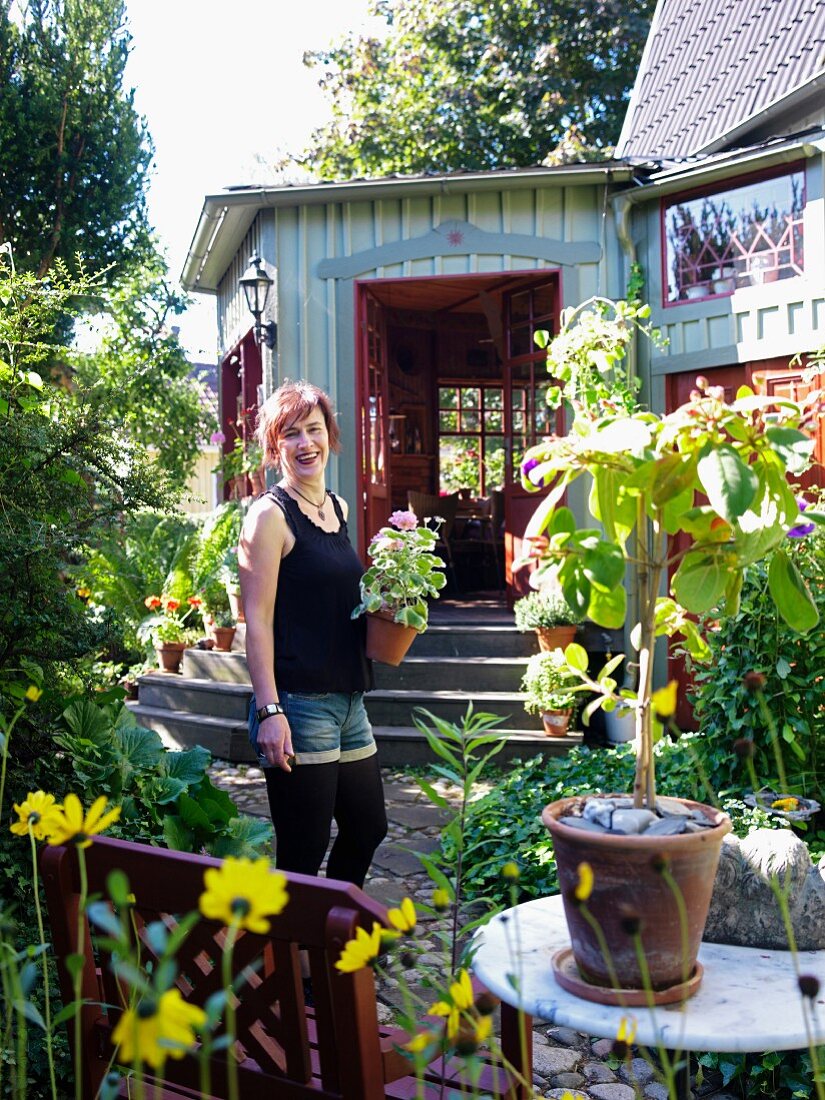 Lachende, junge Frau mit Pflanzentopf in sonnigem Garten vor nostalgischem Holzhaus