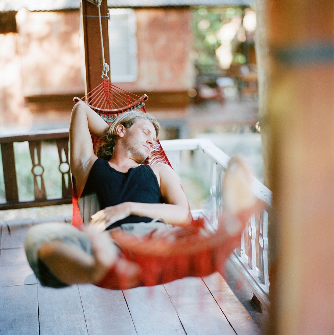 Junger Mann schlafend in roter Hängematte auf Holzveranda
