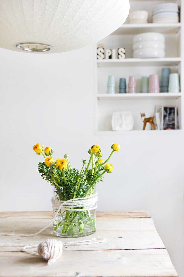 Bouquet of ranunculus in glass vase with decorative cord on wooden table; detail of pendant lamp in foreground and shelving in niche in background