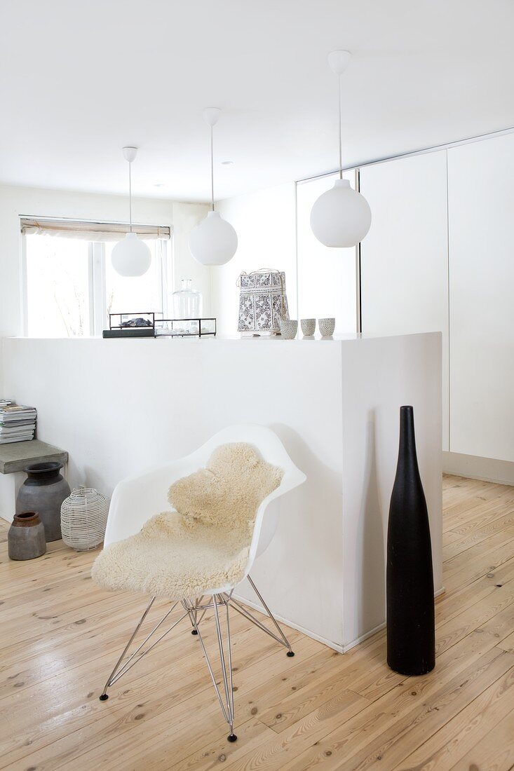 Large, black decorative bottle and classic shell chair with sheepskin rug in front of half-height, masonry wall element and spherical lamps