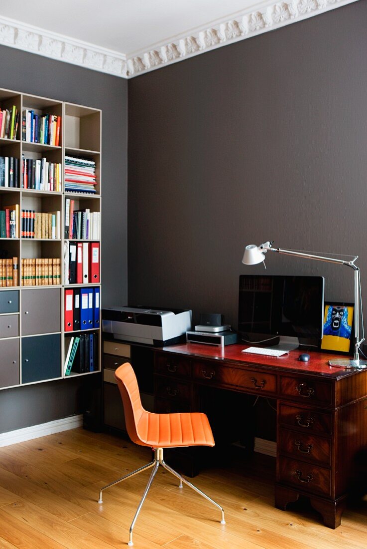 Filigraner Bürostuhl mit oranger Sitzschale vor antikem Sekretär und Designer Tischleuchte in grau getönter Zimmerecke mit Stuckfries, an Wand aufgehängtes Bücherregal