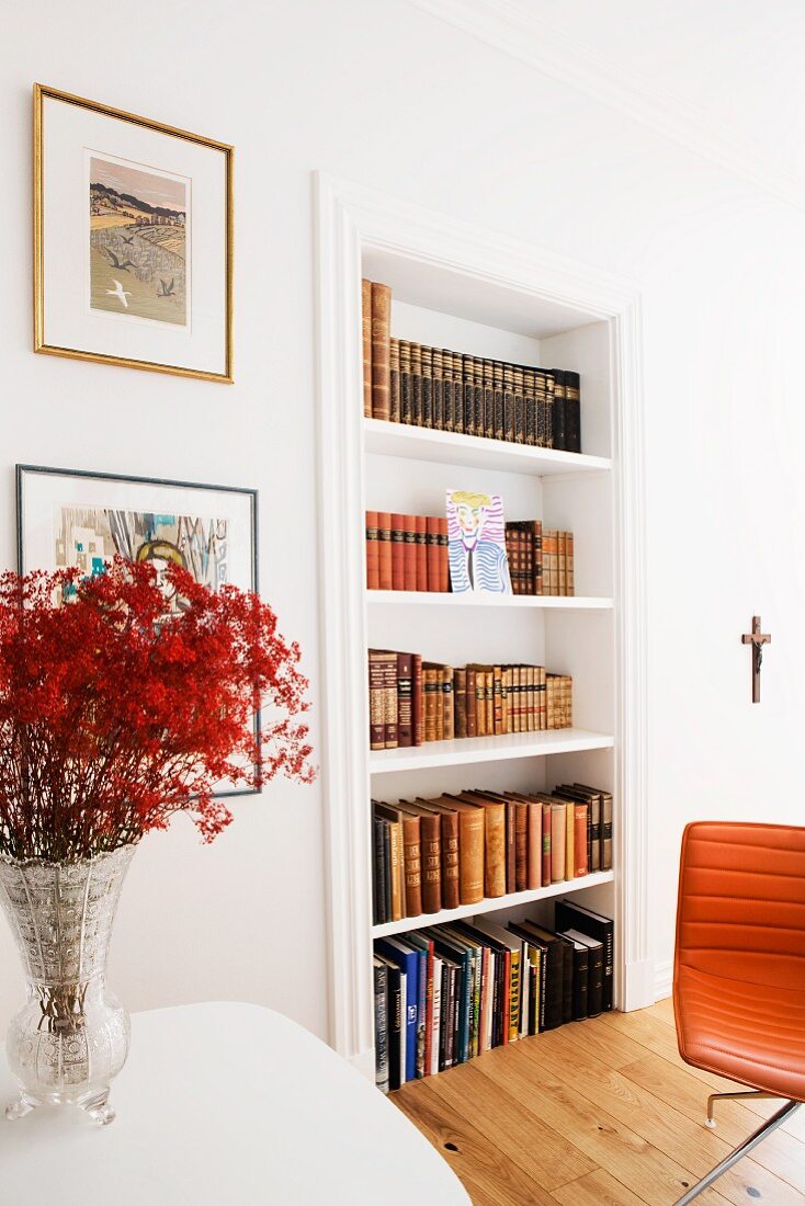 Branches of red flowers in vase next to fitted shelves of antiquarian books and partially visible leather chair