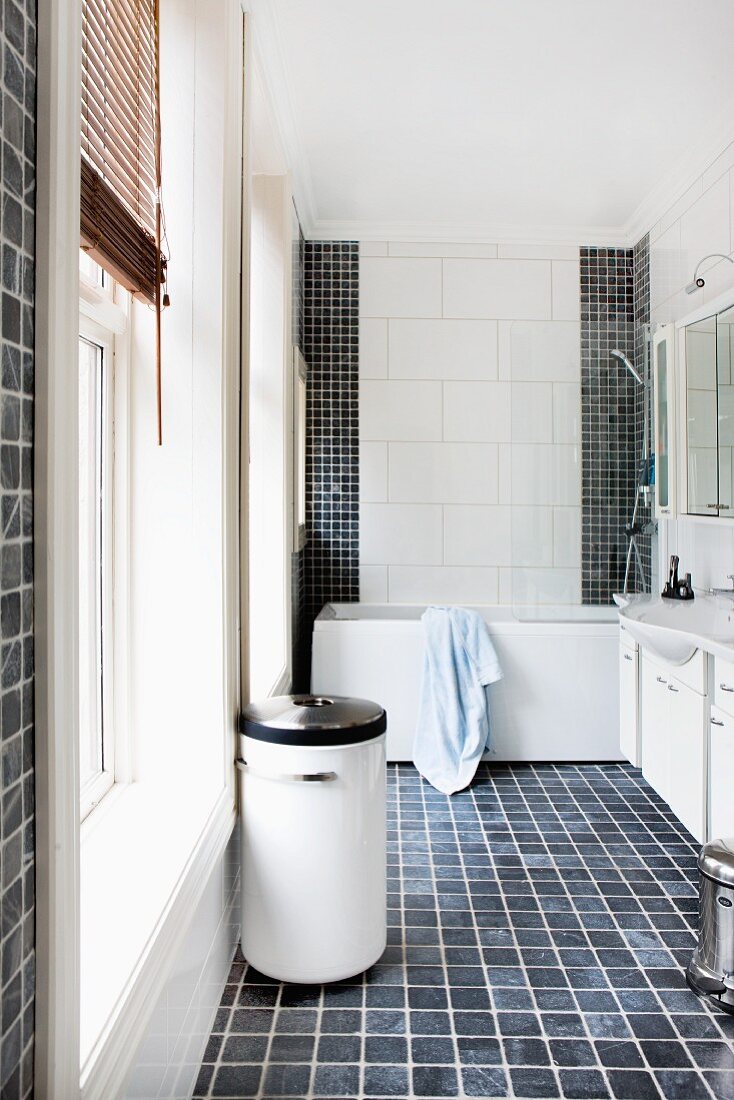 Cylindrical laundry container on dark tiled floor and bathtub in background against wall tiled with tiles of different sizes