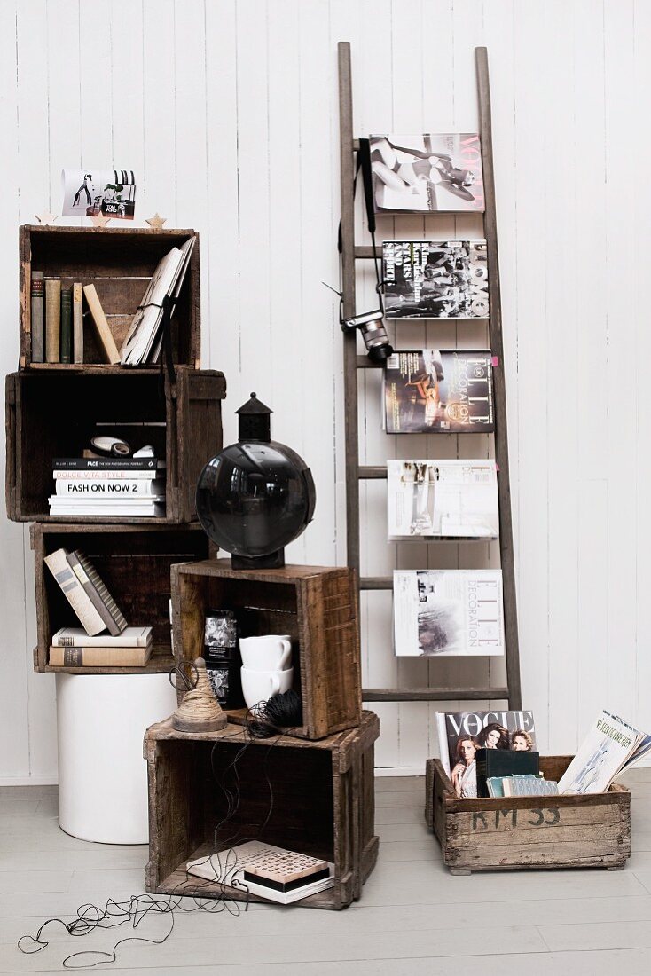 Gestapelte Holzkisten mit Büchern und Deko, daneben eine Leiter mit quergehängten Modezeitschriften