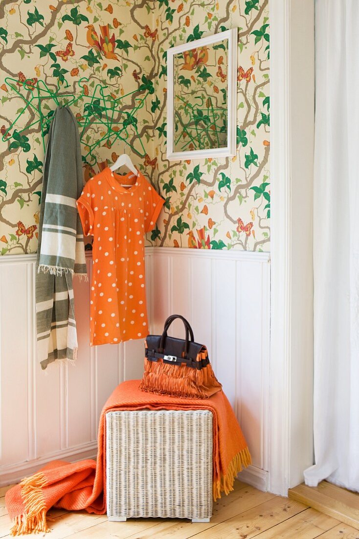 Schlafzimmerecke mit grün floral gemusterter Tapete und Korbhocker vor brüstungshoher Wandvertäfelung; Kleid und Fransendecke in Orange als farbliche Ergänzung