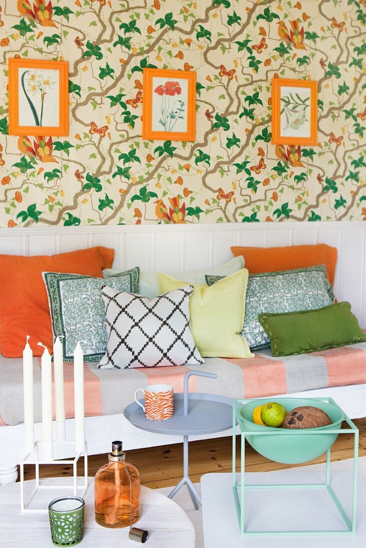 Bench with seat cushion and scatters cushions against white wall panelling below orange-framed botanical pictures on floral wallpaper
