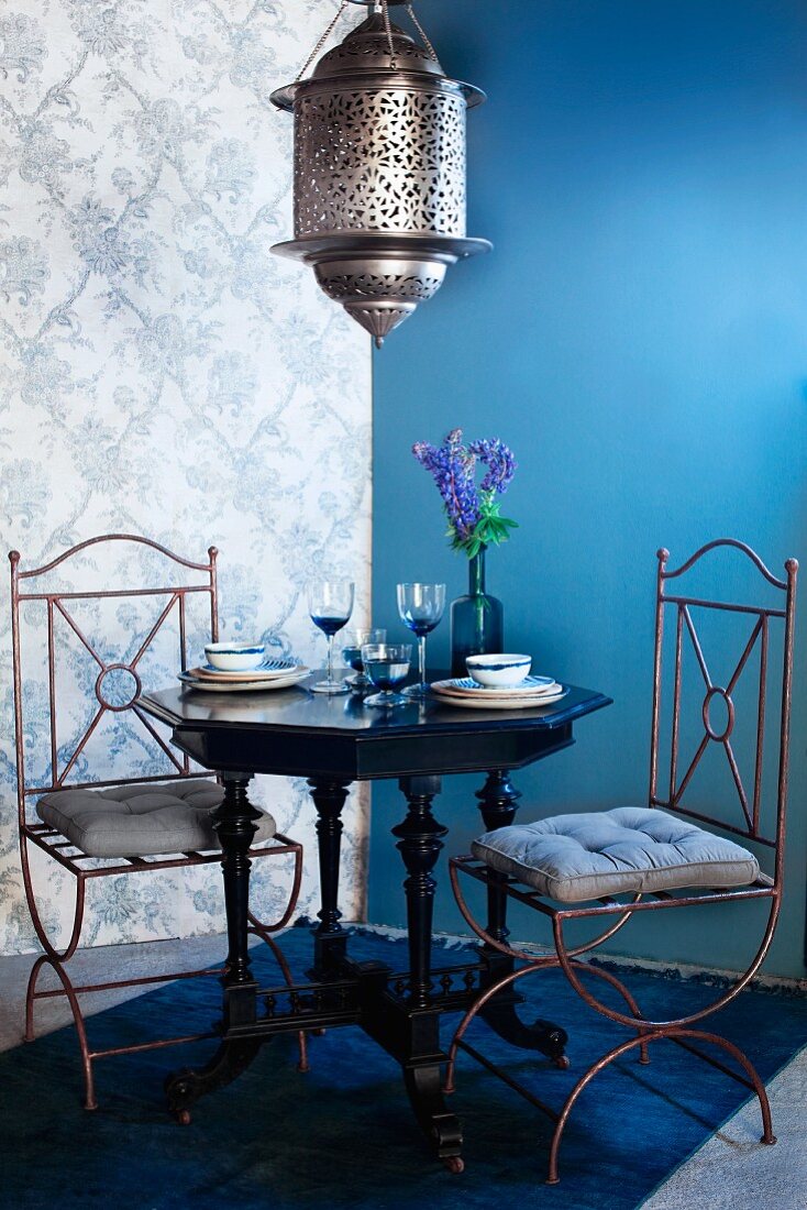 Hängeleuchte im marokkanischen Stil über antikem Tisch mit zwei Drahtstühlen in Zimmerecke mit blauer Wand und gemusterter Tapete