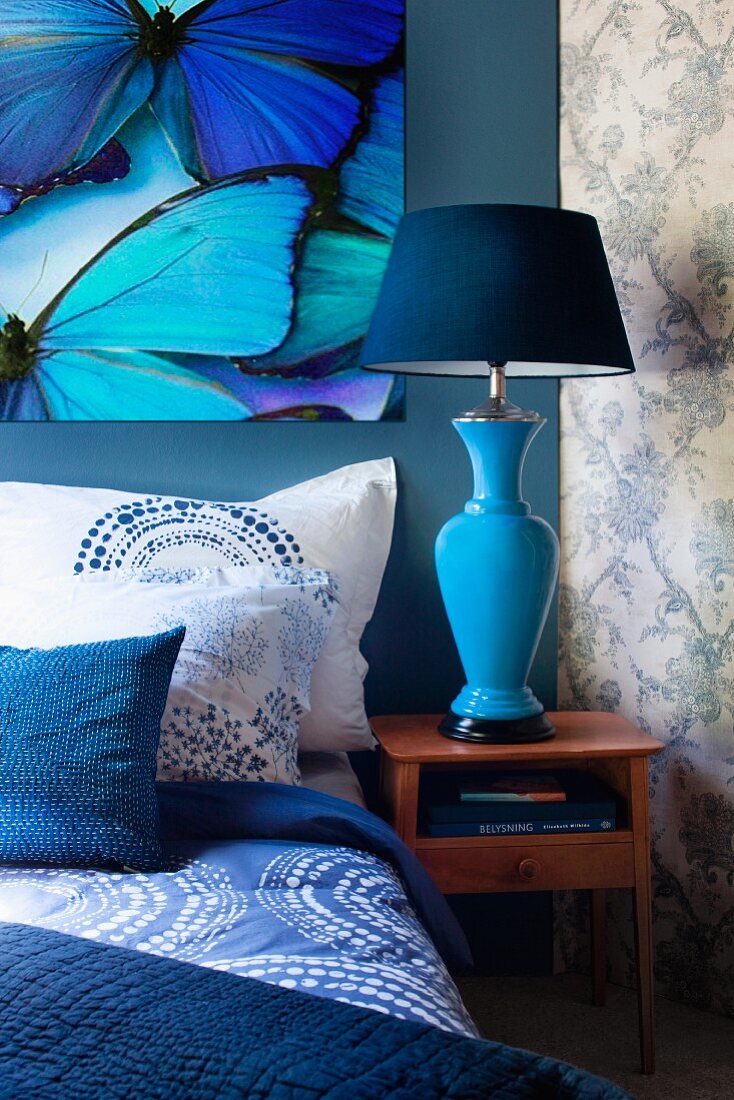 Bett mit Decken und Kissen in Blautönen, darüber Bild mit Schmetterlingen; blaue Nachttischlampe auf Schränkchen