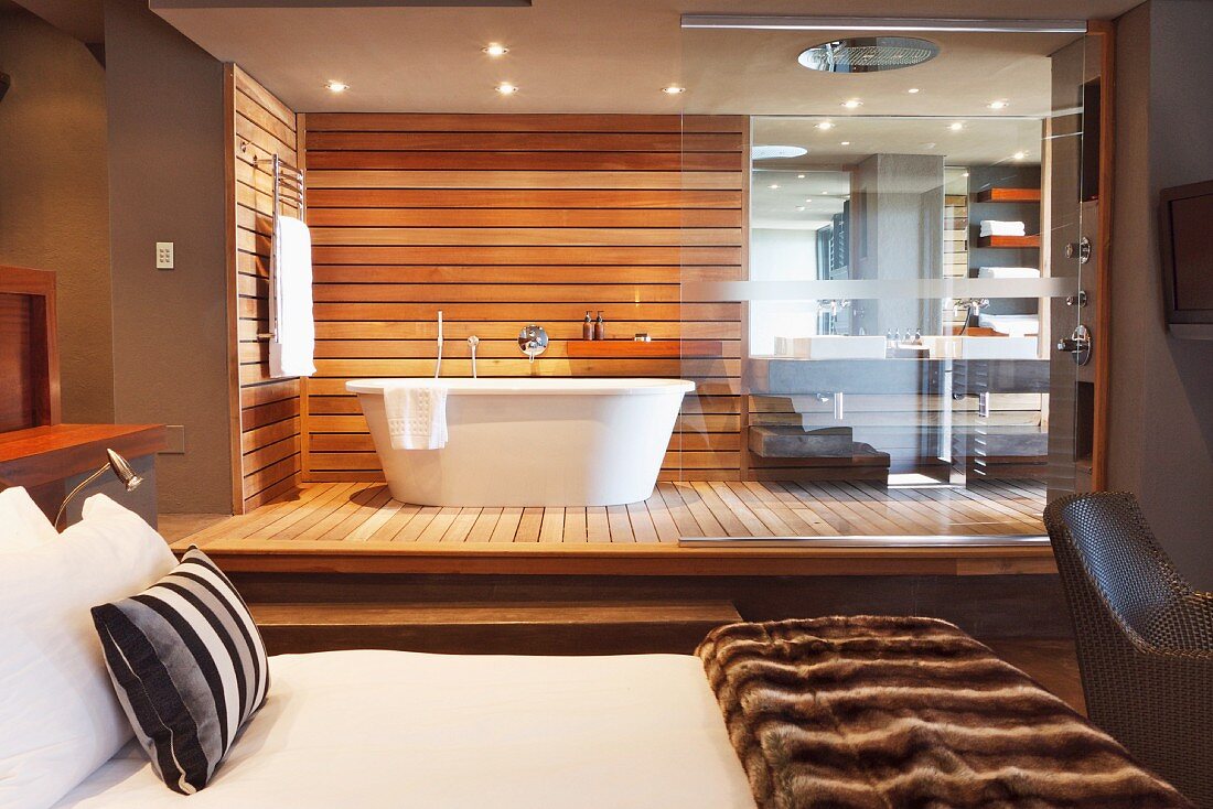Vom Schlafzimmer aus zugängliches Designerbadezimmer mit freistehender weißer Badewanne vor holzverkleideter Wand