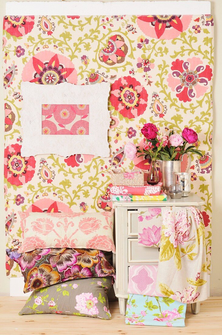 Kissenstapel mit floralen Kissenbezügen vor pastell gemusterter Wandbespannung und Bilderrahmen, daneben Vintagekästchen mit Stoffbahnen drapiert