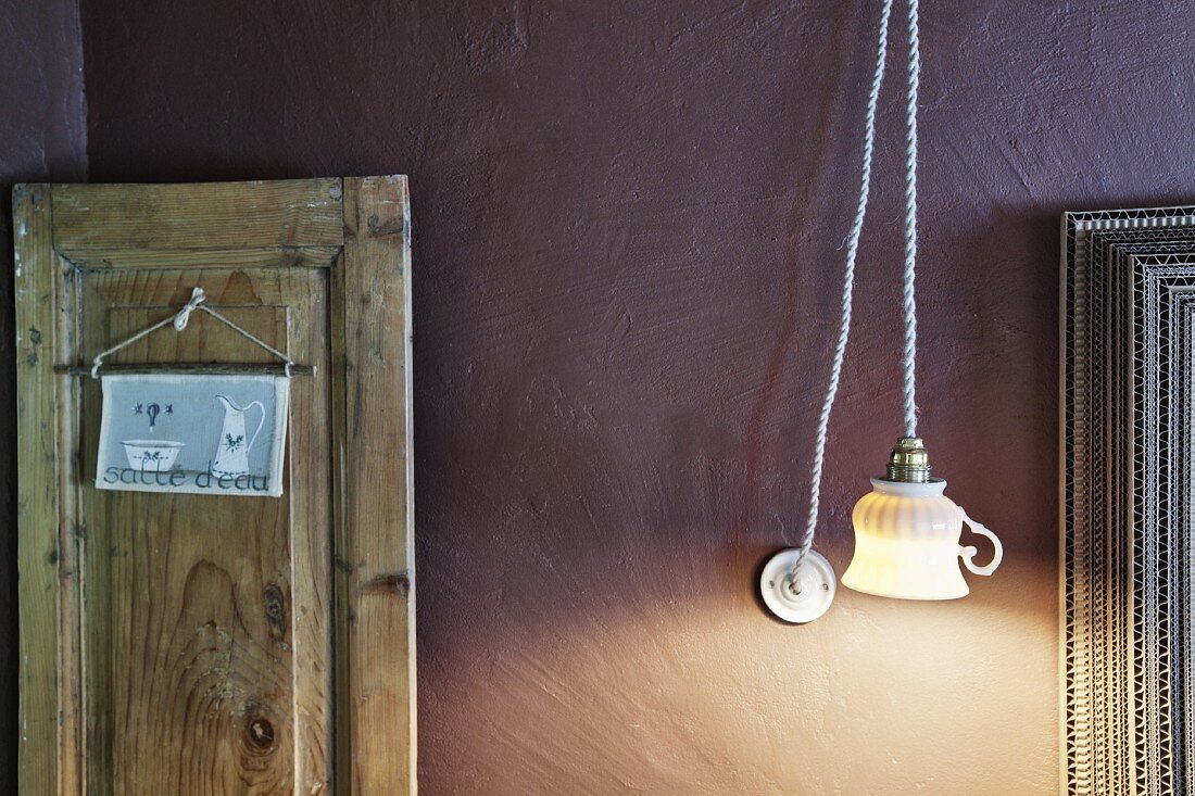 Hängeleuchte mit ehemaliger Tasse als Lampenschirm vor mauvefarbener Wand, Schild an rustikalem Holzpaneel aufgehängt