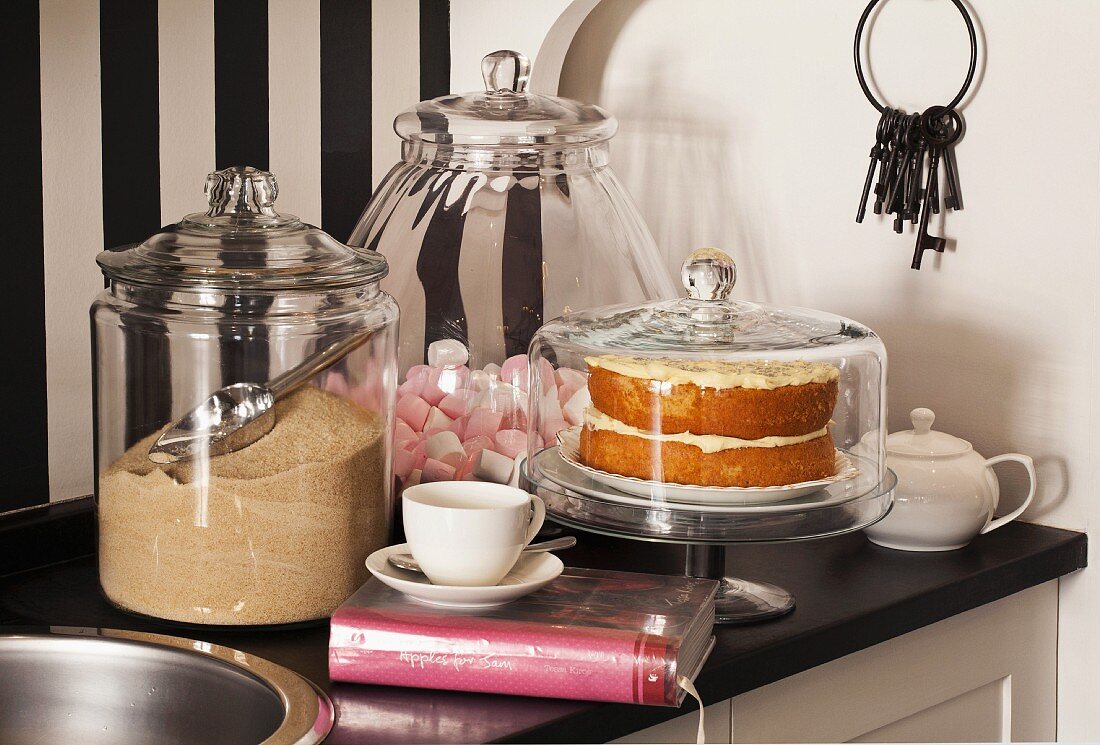 Brauner Zucker und Marschmallows in Glasgefäßen und Kuchen unter Glashaube auf schwarzer Küchenarbeitsplatte