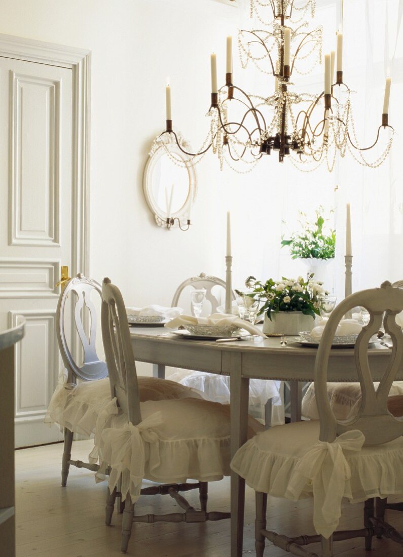 White dining room with festively set table, chandelier & chairs with romantic, flounced seat cushions
