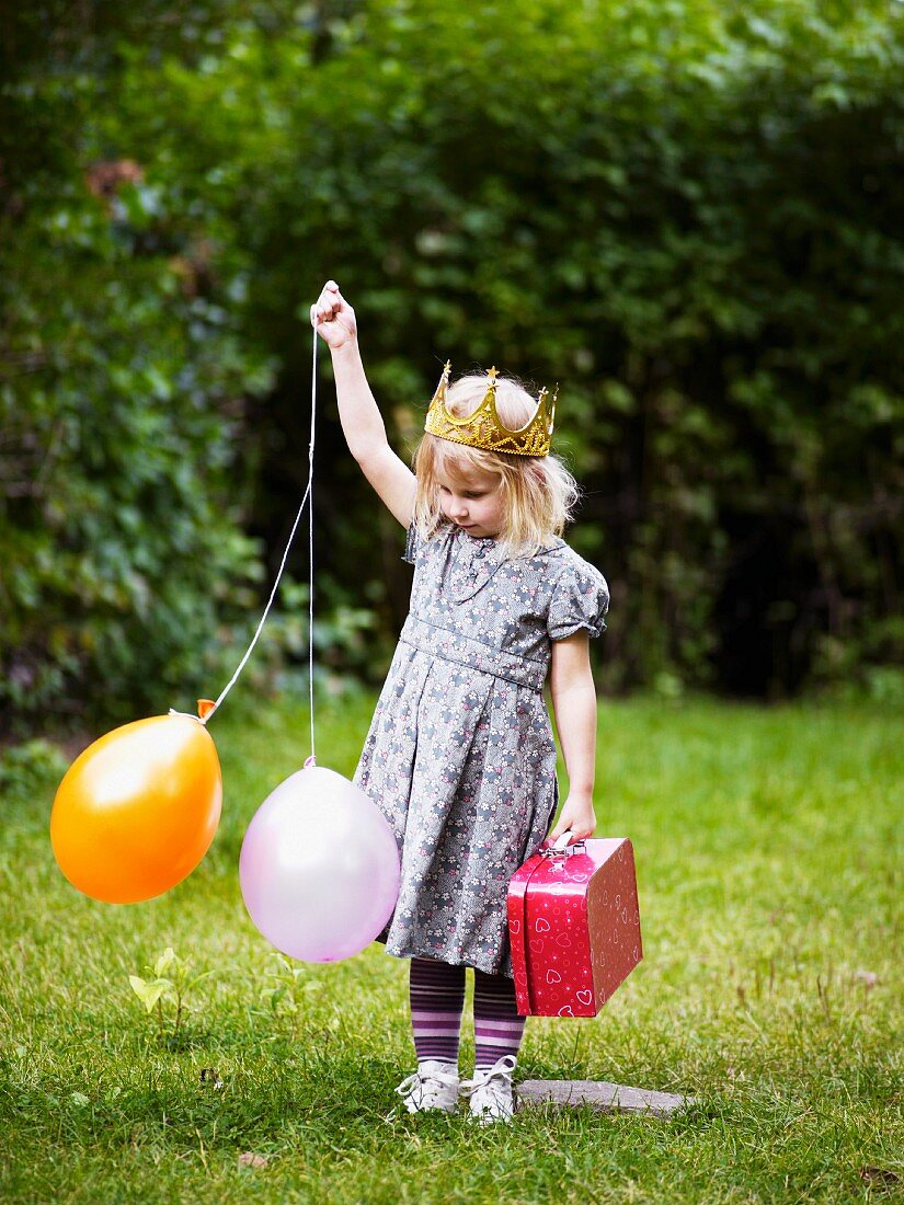 Kleines Mädchen mit Krone, Luftballons & Köfferchen im Garten
