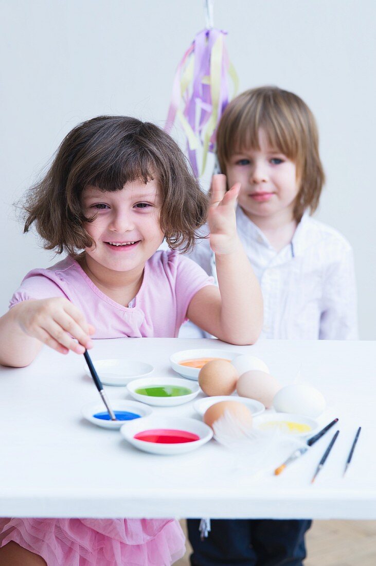 Lachendes Mädchen mit Pinsel und Farben; im Hintergrund ein lächelndes Mädchen mit Spielzeug