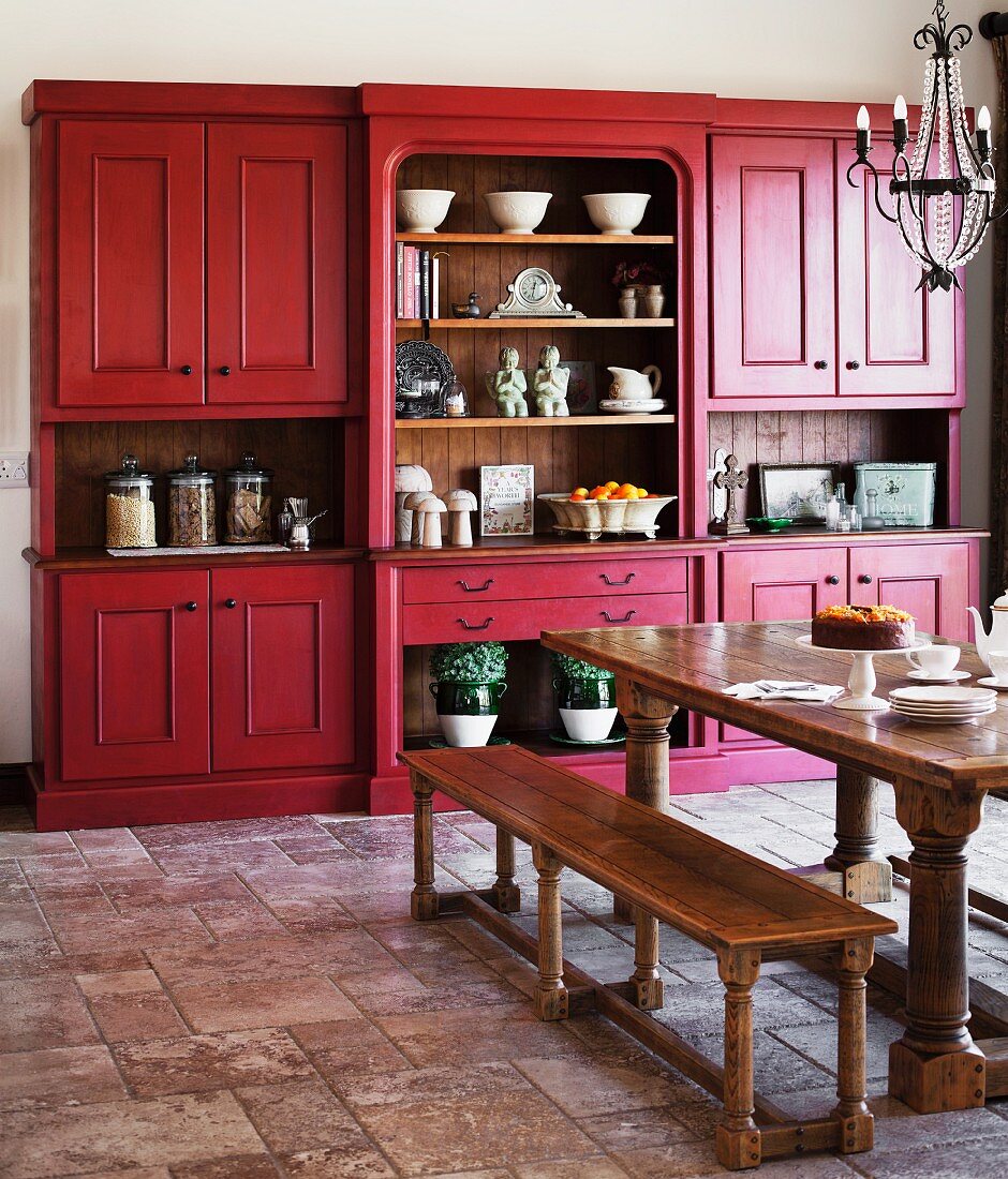 Country-house style kitchen-dining room with heavy wooden table and benches below wrought iron chandelier; matt red kitchen dresser in background