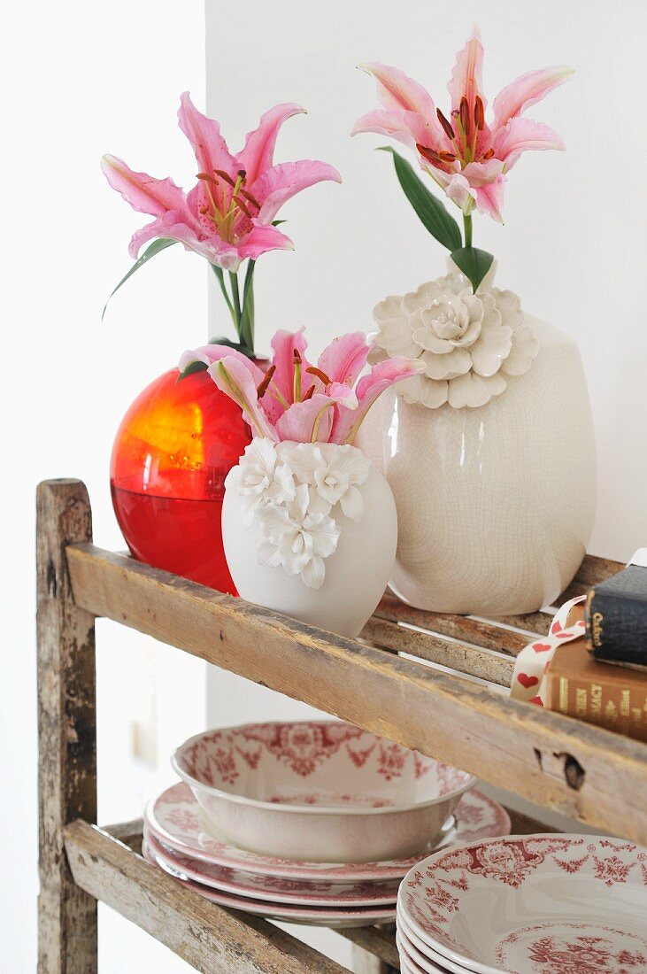 Pink lilies in white china vases and red glass vase next to traditional dinner service on vintage wooden shelving
