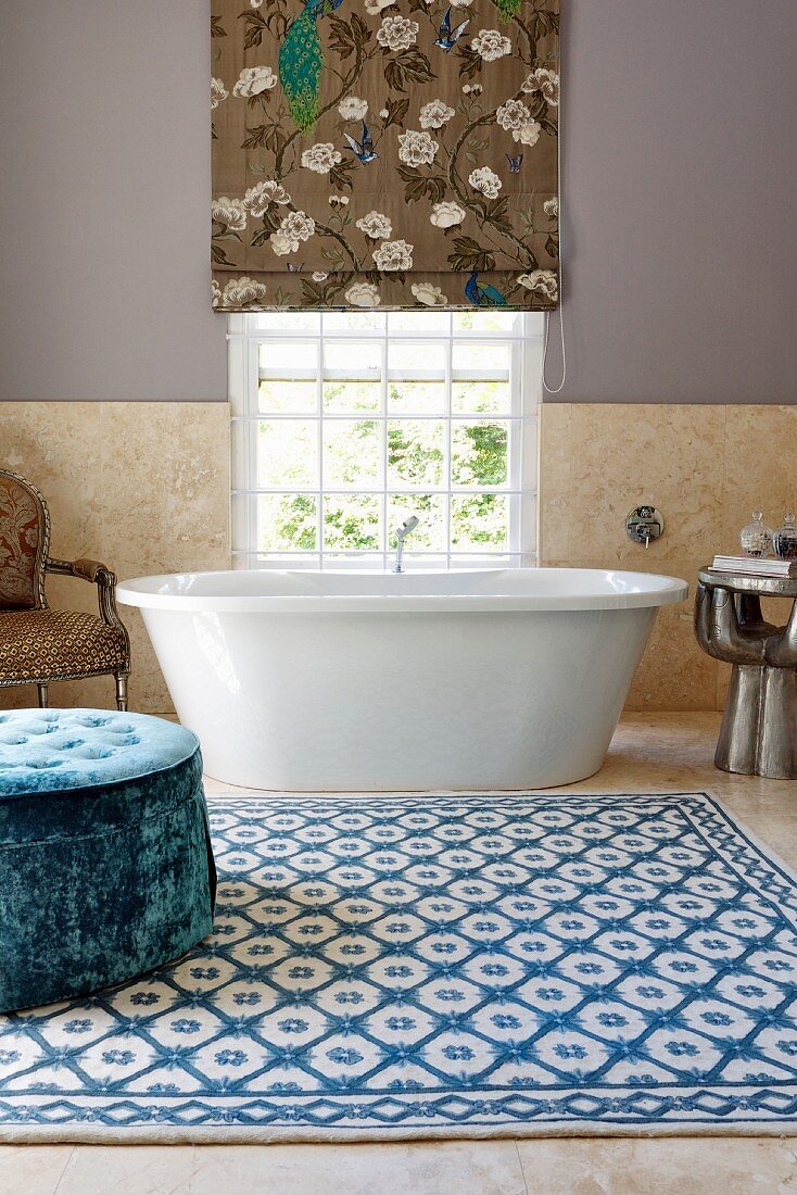 Grossräumiges Bad mit blauem Samt-Polsterhocker auf gemustertem Teppich vor Badewanne am Fenster