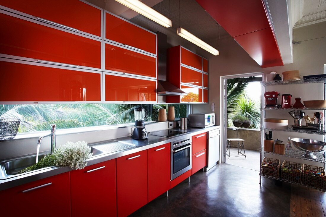Fitted kitchen with red units and metal shelving next to open terrace door