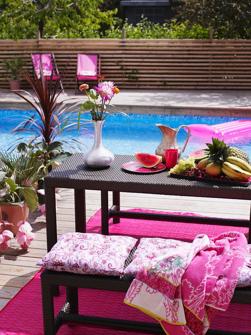 Azurblauer Swimmingpool; im Vordergrund eine Holzterrasse mit pinkfarbenem Teppich und Rattanmöbeln