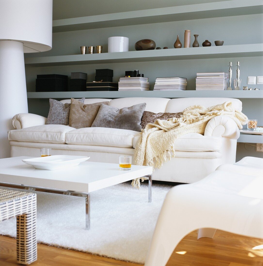 Modernes Wohnzimmer mit grauem Wandregal und überdimensionaler Stehlampe; davor eine weiße Couch mit weißem Couchtisch