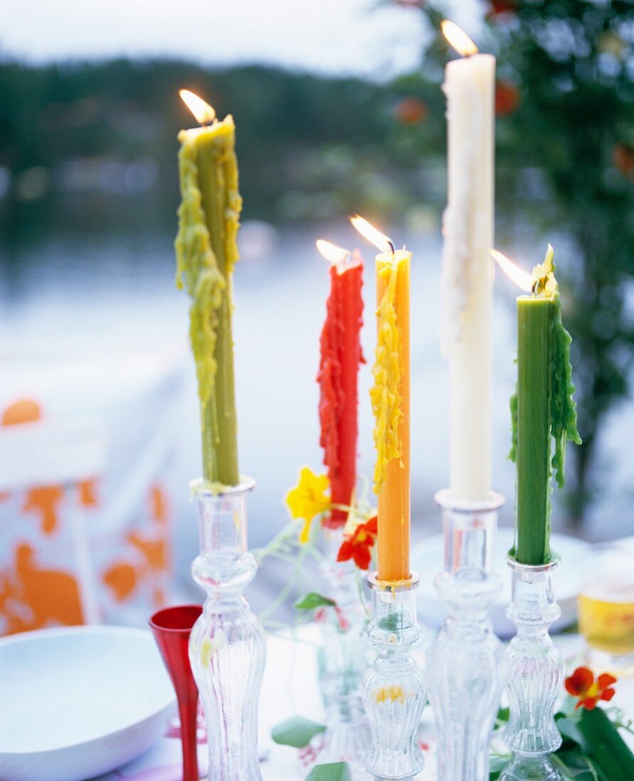 Candles of different colours in glass candlesticks on table in garden