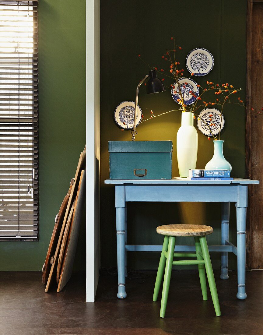 Pastellblauer Tisch mit Hocker an olivgrüner Wand; an der Wand eine Porzellantellersammlung