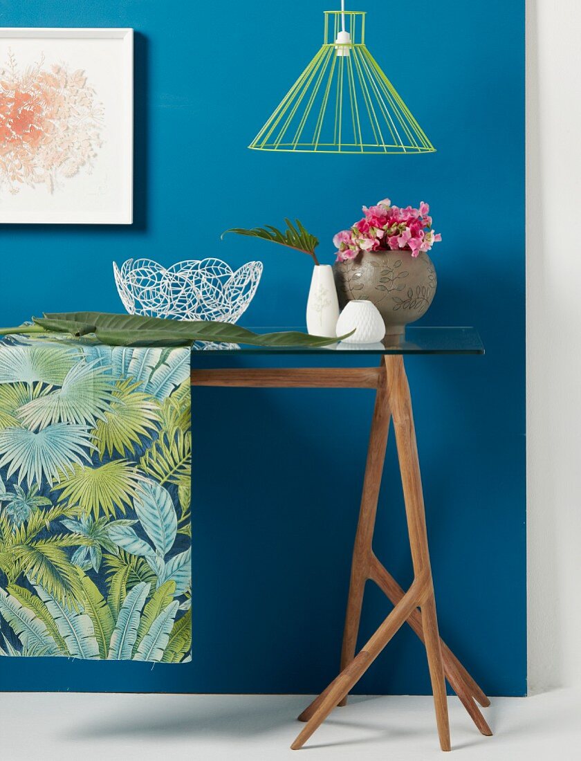 Arrangement auf Glastisch und Tischläufer mit Dschungelmotiv vor blauer Wand