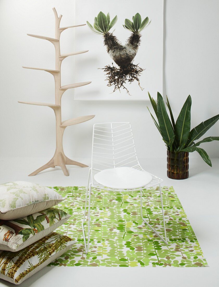 Filigraner weißer Metallstuhl auf Teppich mit Dschungelmotiv und Kissenstapel, im Hintergrund naturbelassener, kunsthandwerklicher Garderobenbaum