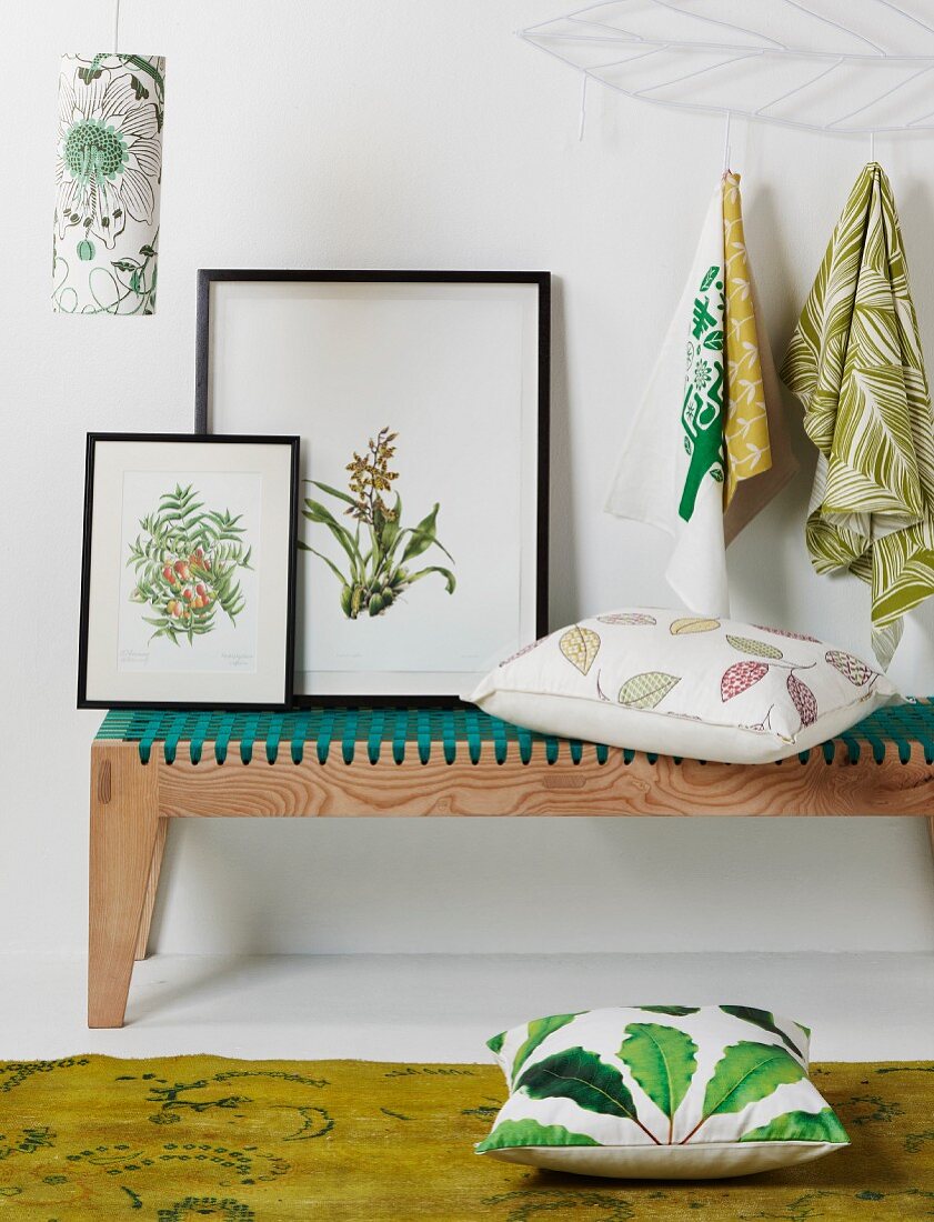Botanical drawings leaning against wall on decorative, modern wooden bench and scatter cushions below patterned cloths hanging from hooks