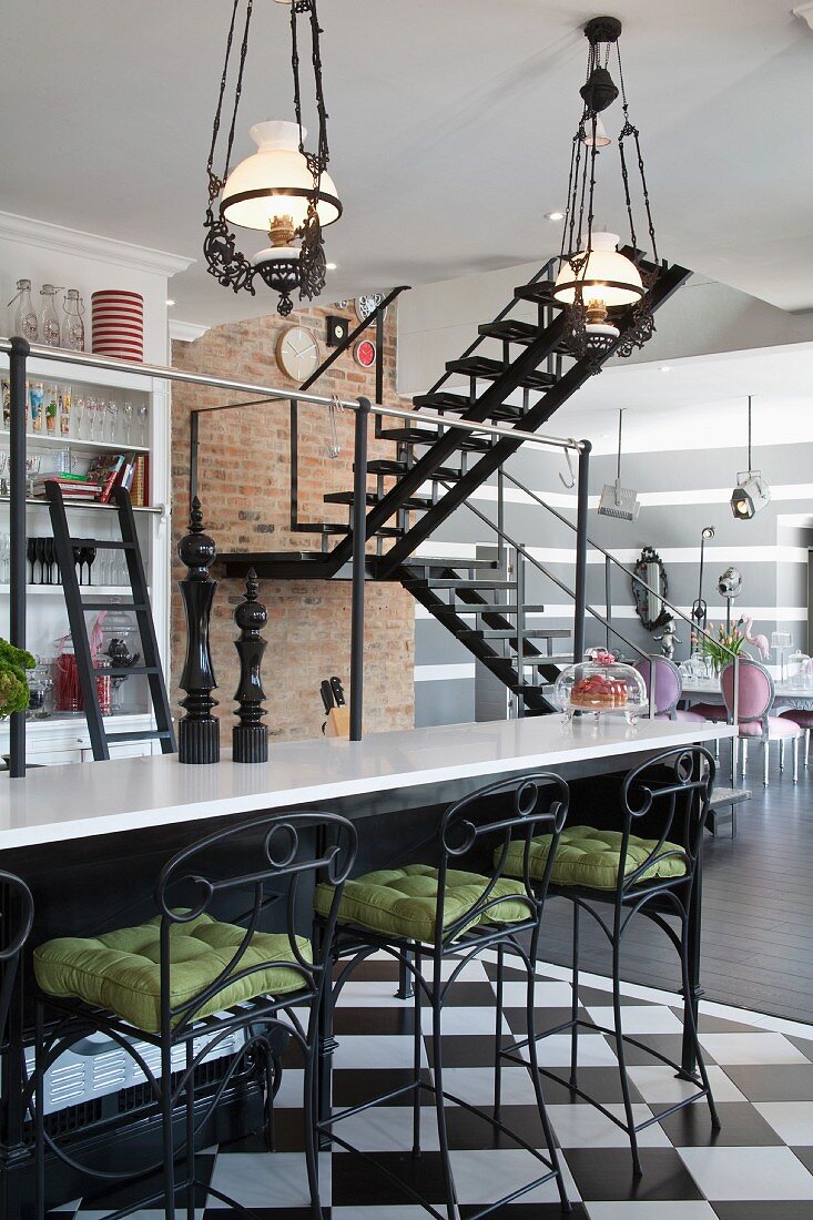 Bar stools with wrought iron and green seat cushions in front of breakfast bar under vintage, pendant gas lamps and staircase in background in open-plan interior