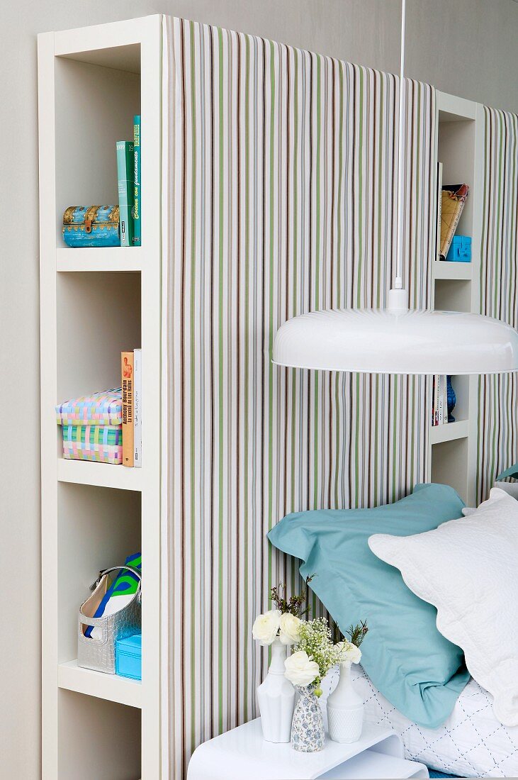 Pendant lamp with white lampshade in front of shelving units with striped fabric covers used as headboard