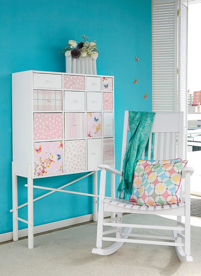 Chest of drawers with patchwork decoupage drawer fronts against blue wall and white wooden rocking chair