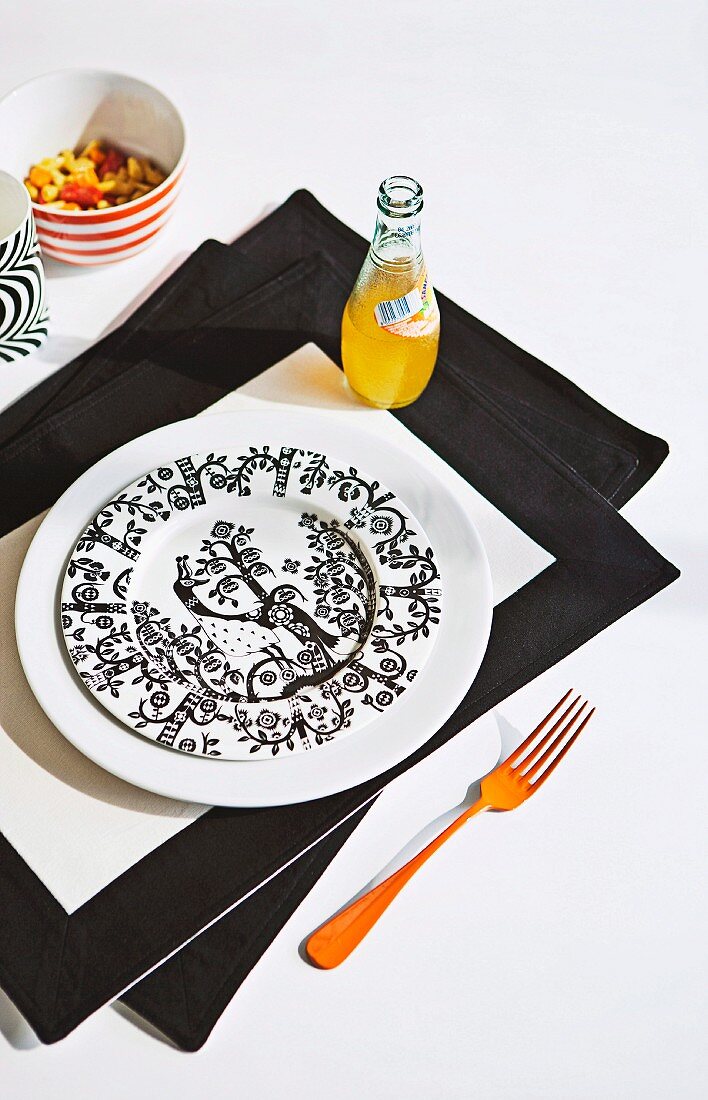Schwarzweiss gemusterter Teller und Limonadenflasche auf schwarzweissem Tischset, an der Seite orange gefärbte Gabel