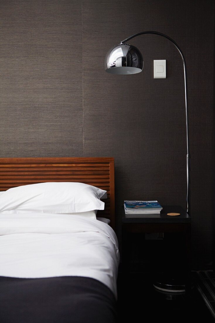 Purist double bed, shiny standard lamp and black bedside table against grey, structured wallpaper