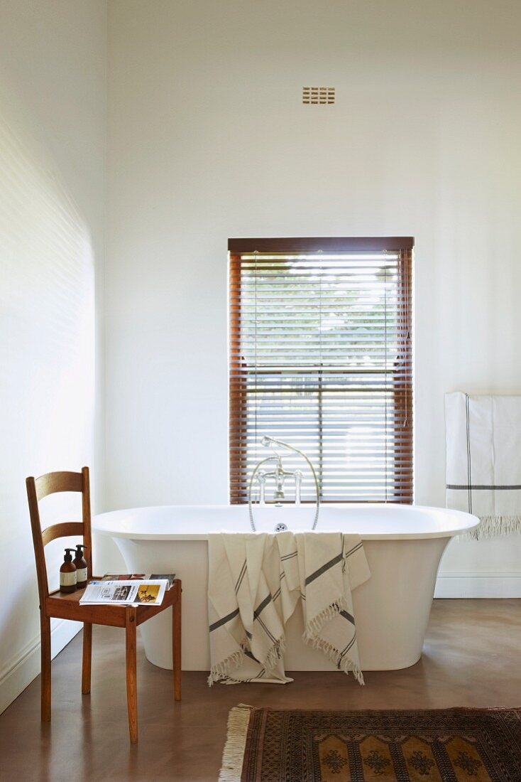 Towel draped over free-standing bathtub below window with closed louver blind, toiletries on simple wooden kitchen chair to one side