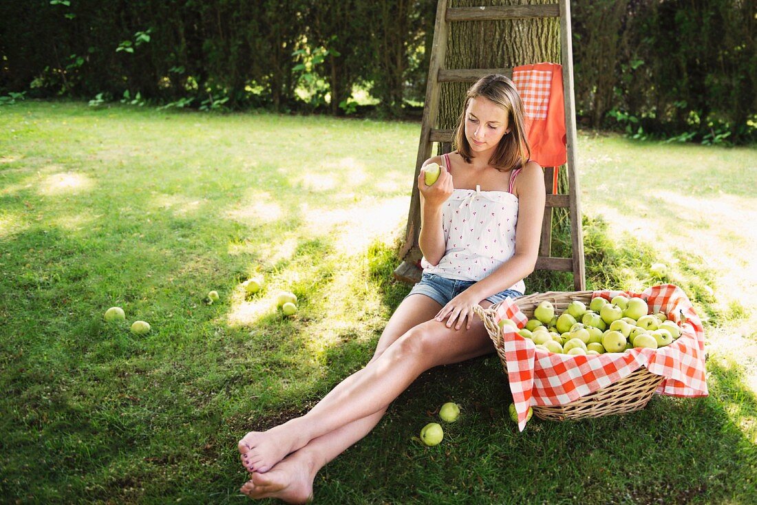 Junge Frau mit Apfel in der Hand neben Apfelkorb am Baum sitzend