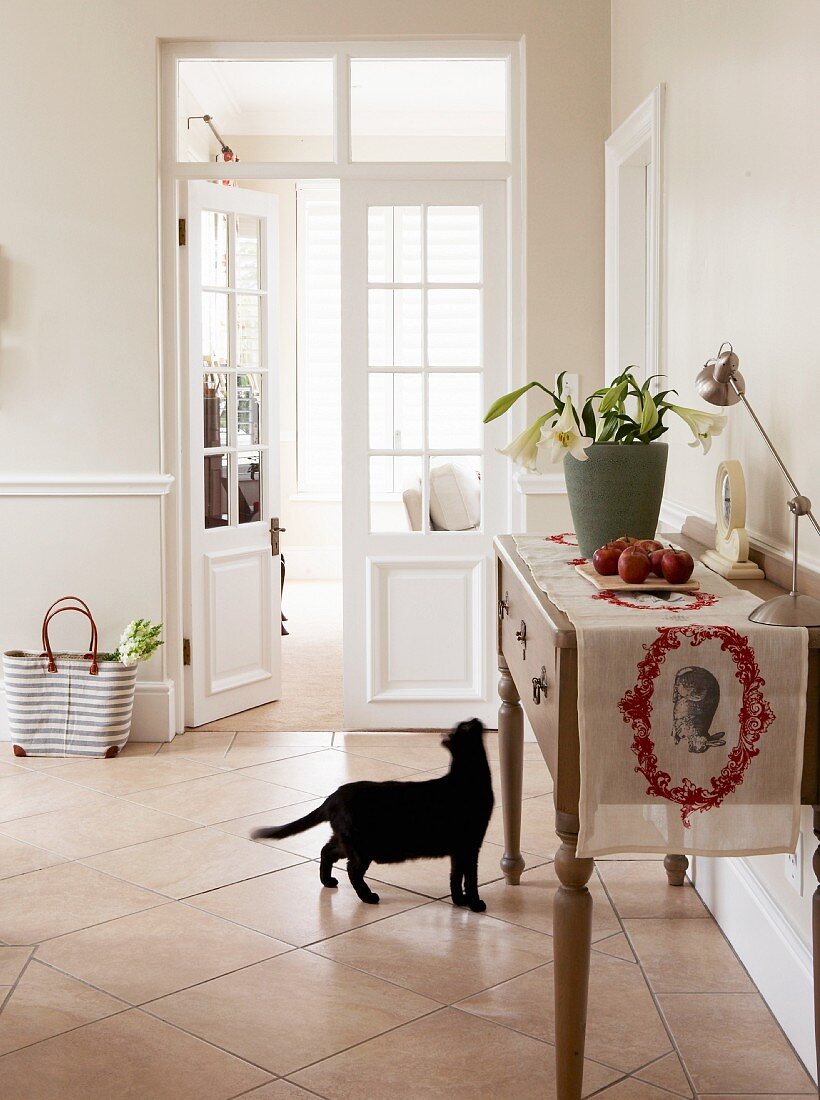 Schwarze Katze vor Sideboard mit Blumendeko und Tischläufer in einer hellen Landhausküche