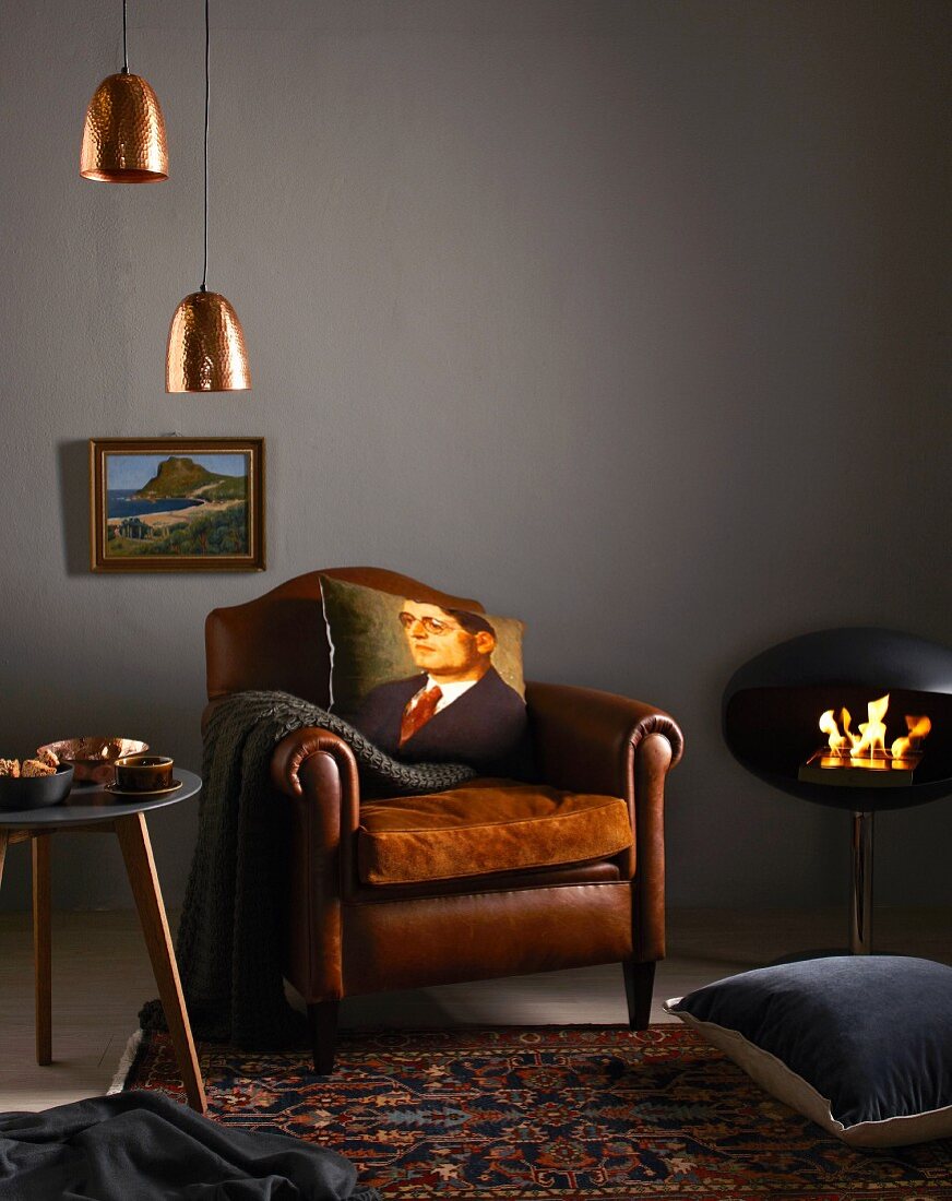 Cushion printed with portrait of man on leather armchair, copper lampshades and modern fireplace