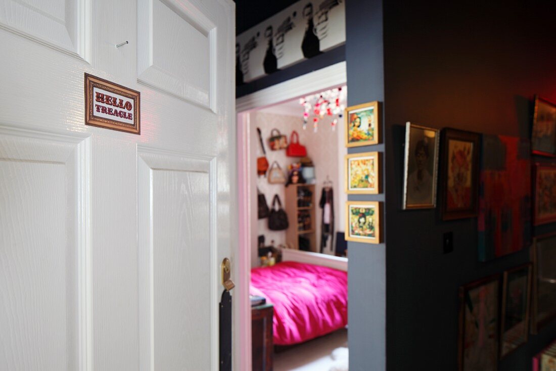 View into feminine bedroom with white-painted panelled door and gallery of pictures in dark hallway