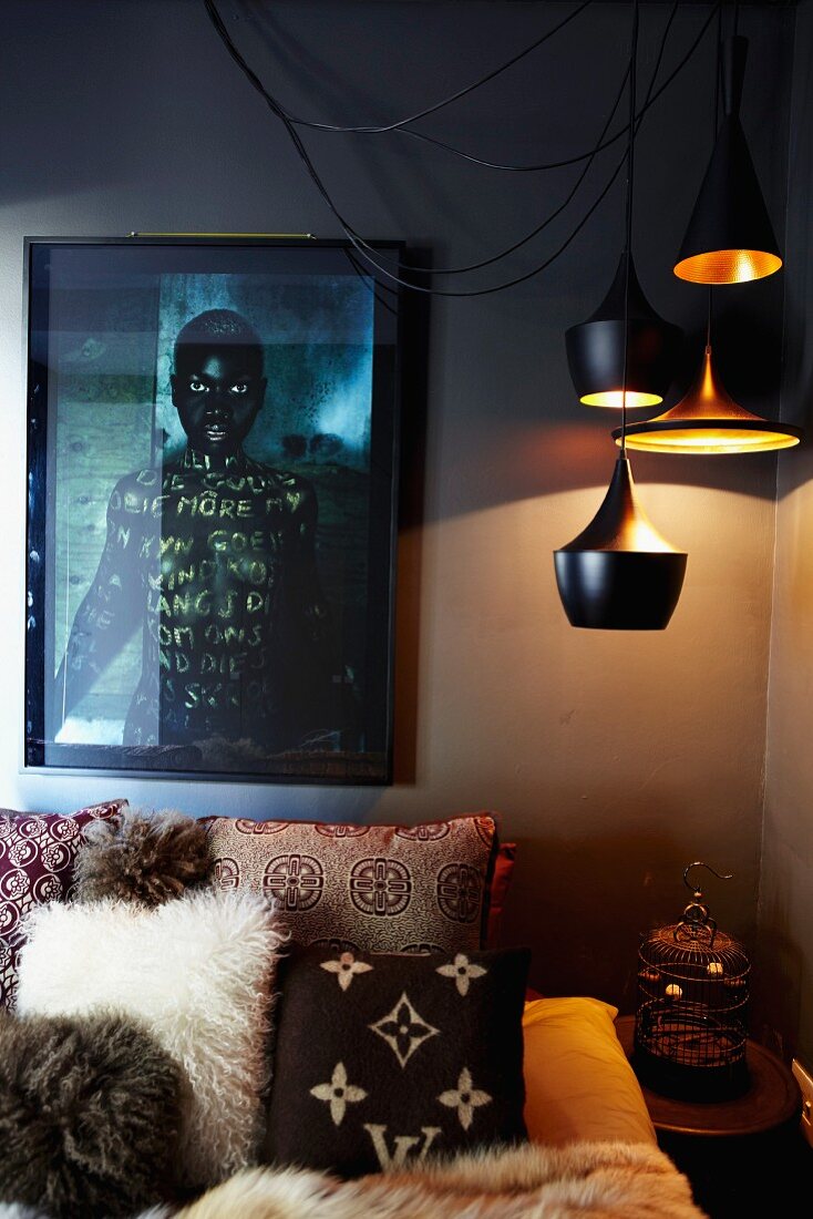 Dramatische Beleuchtung in Schlafzimmerecke - Verschiedene Pendelleuchten über Kissen mit folkloristischen Mustern auf Bett, vor dunkel getönter Wand und Photographie eines dunkelhäutigen Menschen