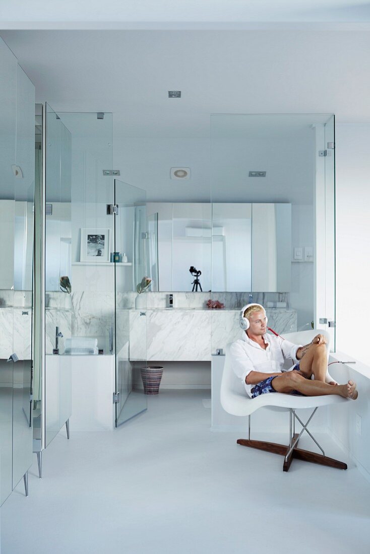 Mann auf Klassiker Stuhl La Chaise, vor modernem, verglastem Badbereich mit Marmor Waschtisch und mehrteiligem Spiegelschrank