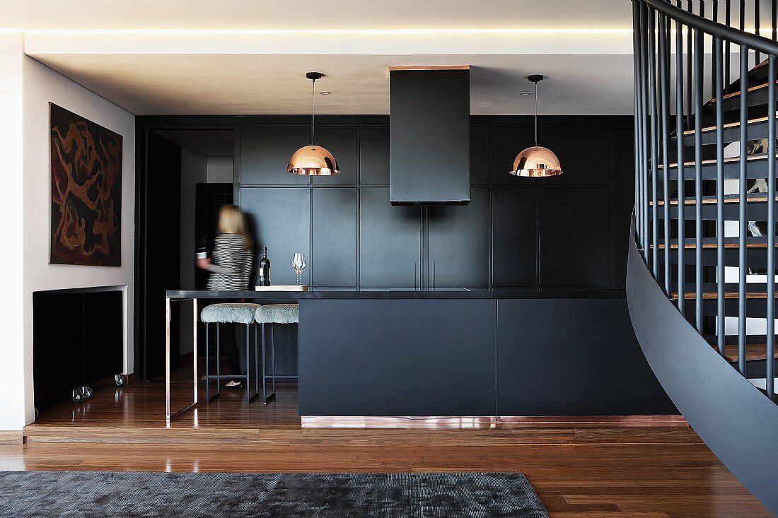 Open-plan, black kitchen and pendant lamps with copper-coloured lampshades in interior with partially visible spiral staircase