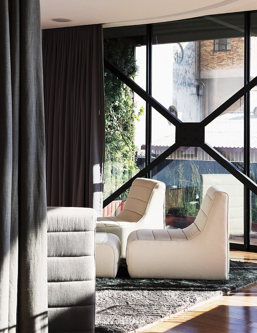 White easy chairs on rug in front of glass facade with dark, floor-length curtains