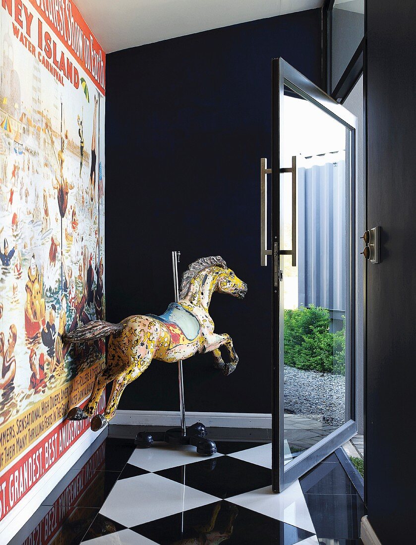 Old carousel horse next to wall with vintage poster in entrance hall with chequered floor and glass door