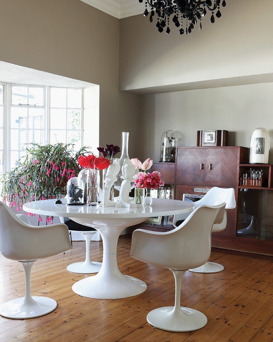 weiße Klassiker Drehstühle um runden Tisch mit Blumenvasen, im Hintergrund Blumen vor Terrassen Sprossentüren, in grau getöntem Wohnzimmer mit schlichtem Dielenboden