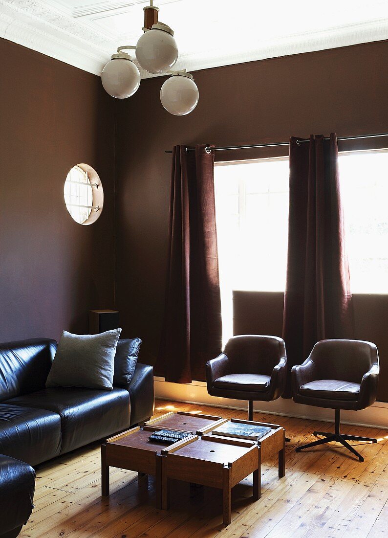 Elegant living room with walls painted dark brown, black leather sofa and swivel chairs around wooden coffee table and Art Deco ceiling lamp