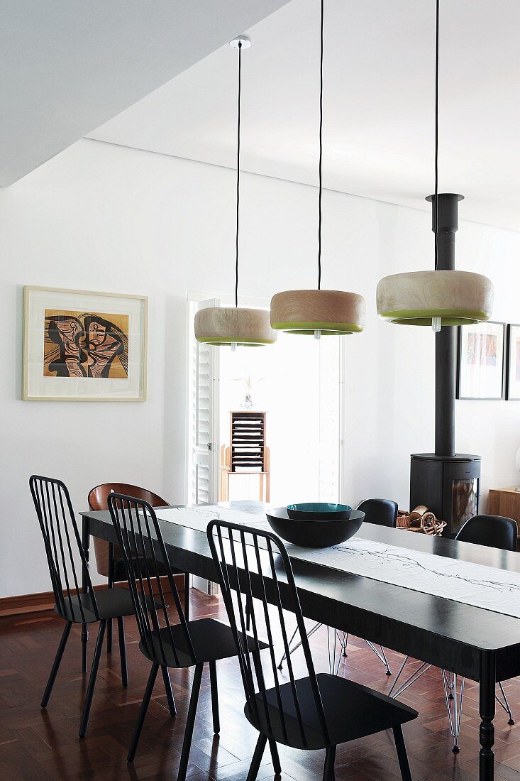 Modern dining room with black dining table below designer pendant lamps; black wood-burning stove in background next to basket of firewood