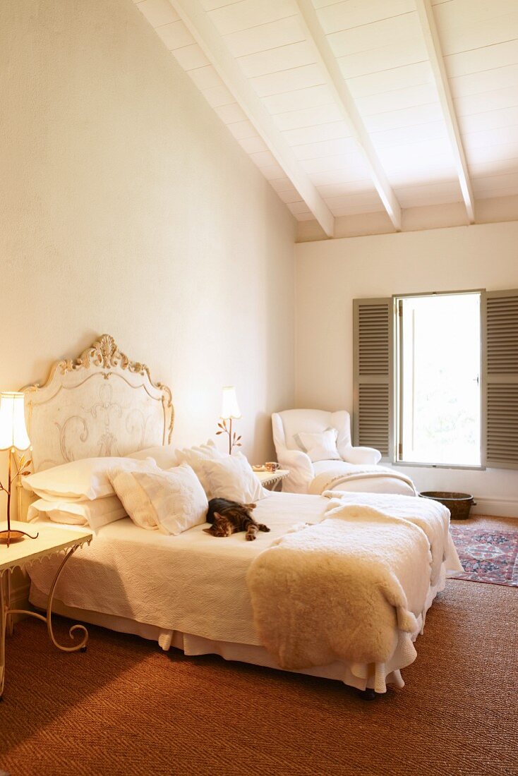 Katze auf Doppelbett mit geschwungenem Kopfteil im Rokoko-Stil in schlichtem Schlafzimmer mit weisser Holzdecke im Dachgeschoss